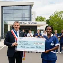 Un maire remet un chèque géant de 30 000 € à une infirmière devant l’entrée de l’Hôpital Central, en présence d’un groupe de soignants applaudissant. La scène se déroule à l’extérieur, sous un ciel clair, le 14 octobre 2024.
